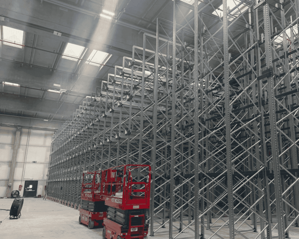 Imponente installazione di scaffalature portapallet industriali in acciaio zincato all'interno di un magazzino logistico, con piattaforme a forbice rosse impiegate per il montaggio.